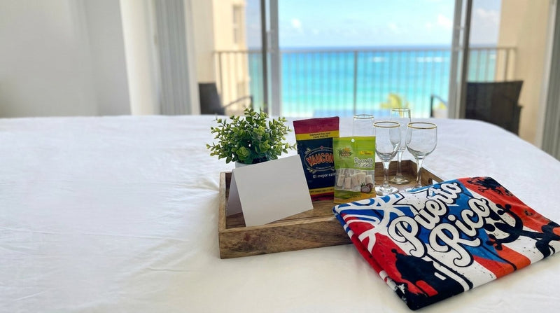 Bandeja de bienvenida para huéspedes con café Yaucono, pilones de coco y copas sobre una cama blanca, con toalla de Puerto Rico y vista al mar